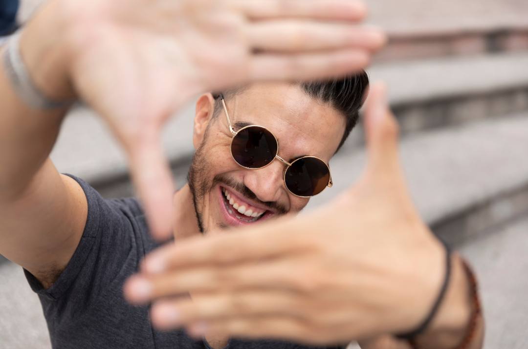 A man wearing glasses is playfully framing his hands in front of his face, looking through them toward the camera with a wide smile and laughing.